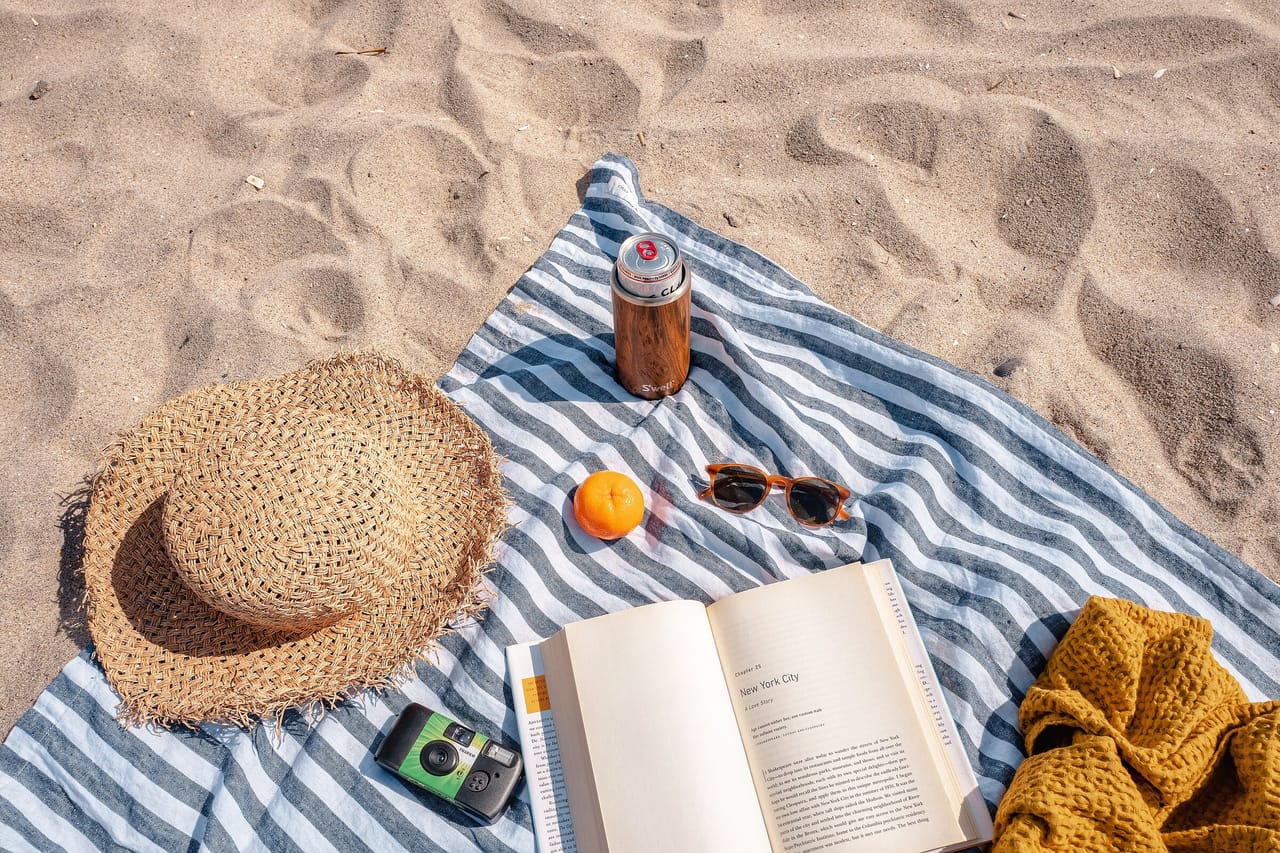 Gestreiftes Strandtuch im Sand mit Strohhut, aufgeschlagenem Buch, Sonnenbrille, Orange und Trinkflasche