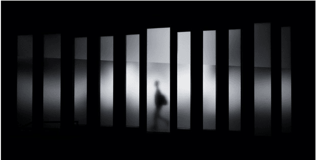 Verschwommene menschliche Silhouette hinter hellen, senkrechten Milchglas-Streifen im Dunkeln