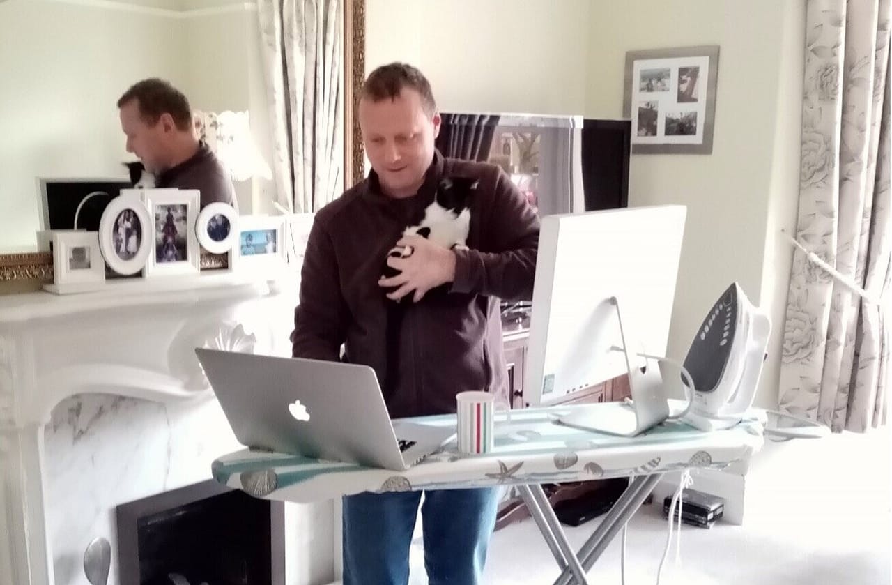 Mann am Bügelbrett als improvisiertem Stehpult, Laptop davor, Katze auf dem Arm, Bügeleisen daneben