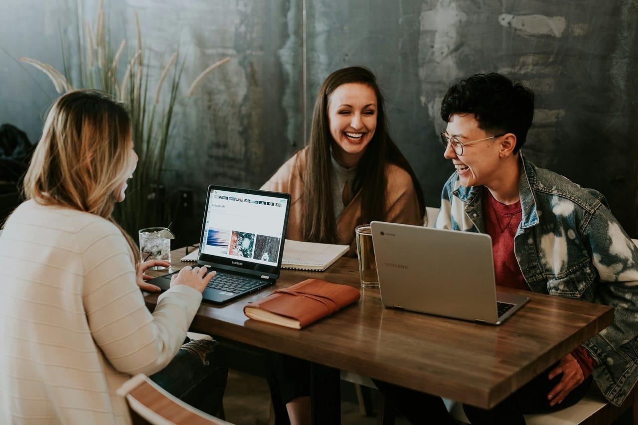 Drei Frauen lachen gemeinsam an einem Holztisch mit zwei Laptops, Notizbuch und Gläsern
